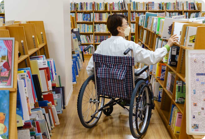 写真:図書館の本棚の間を車椅子で移動する女の人。通路がゆったりしているので、車椅子でも移動しやすいです。