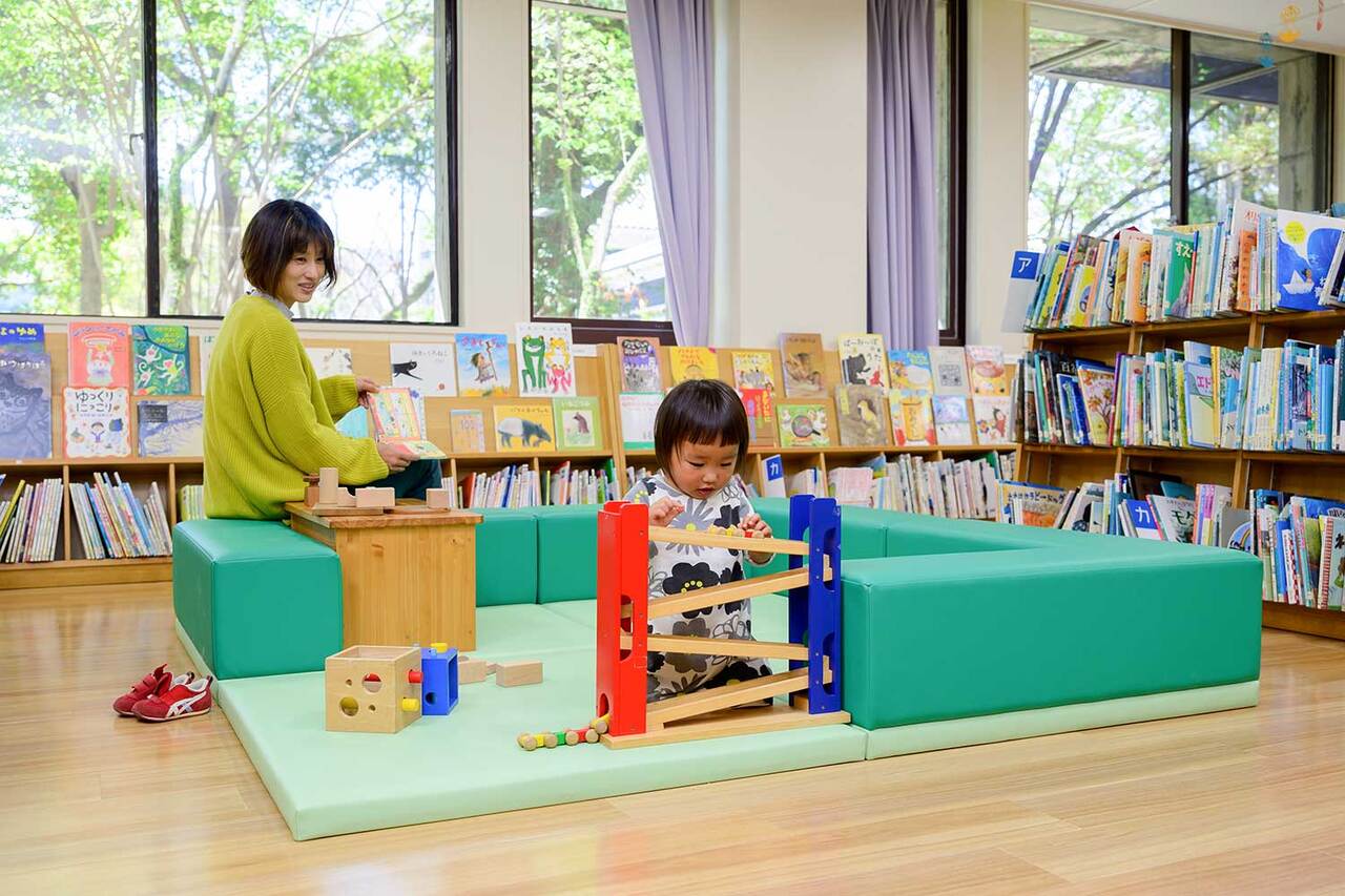 写真:本棚に囲まれた緑色の遊び場で小さな子が遊んでいます。お母さんも近くに座っています。