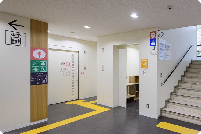写真:建物1階の階段の横に女性用トイレ、多目的トイレ、授乳室があります。床には点字ブロックが敷かれています。