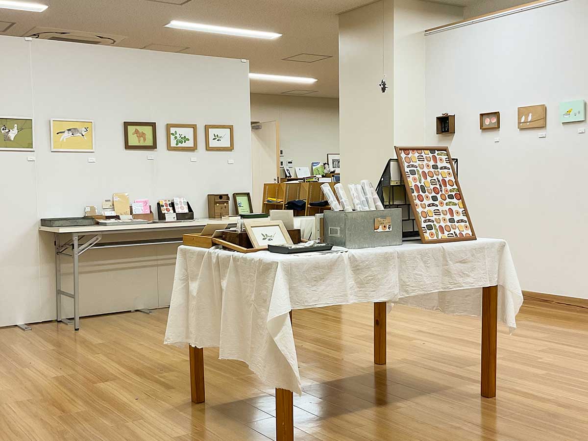 写真：イラスト展の様子。壁にイラストが並んでいます。部屋の真ん中の机にはグッズが展示されています。