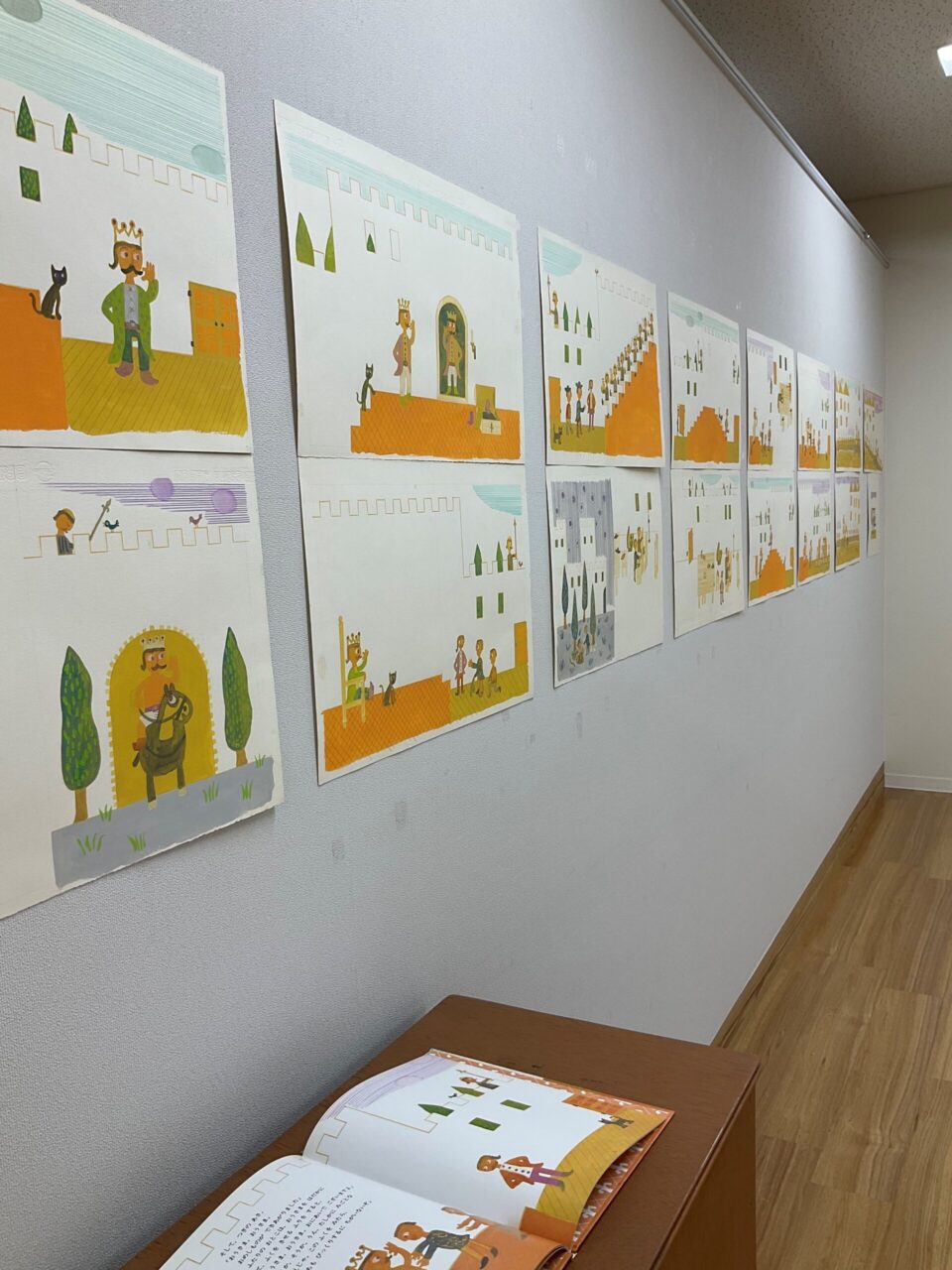 会場の壁に絵が並んでいるようす。「はだかのおうさま」の本と原画。